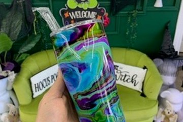 Hand holding a colorful swirl-patterned tumbler with a neon 'Be Creative' sign and green decor in the background.