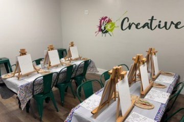 Art workshop setup with easels, canvases, and paint supplies on tables. 'Be Creative' on wall.