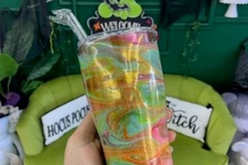 Hand holding colorful tumbler in front of green sofa, neon 'Be Creative' sign overhead.