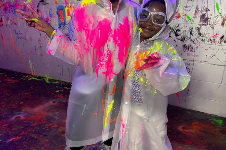 Two kids in goggles and paint-splattered raincoats stand in a graffiti-covered room.